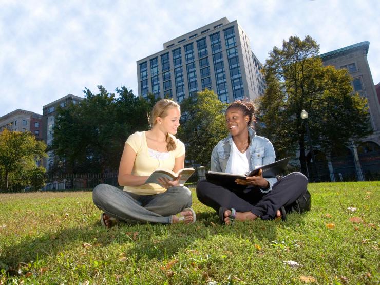 Students sitting on grass and looking at brochures with a building behind them.