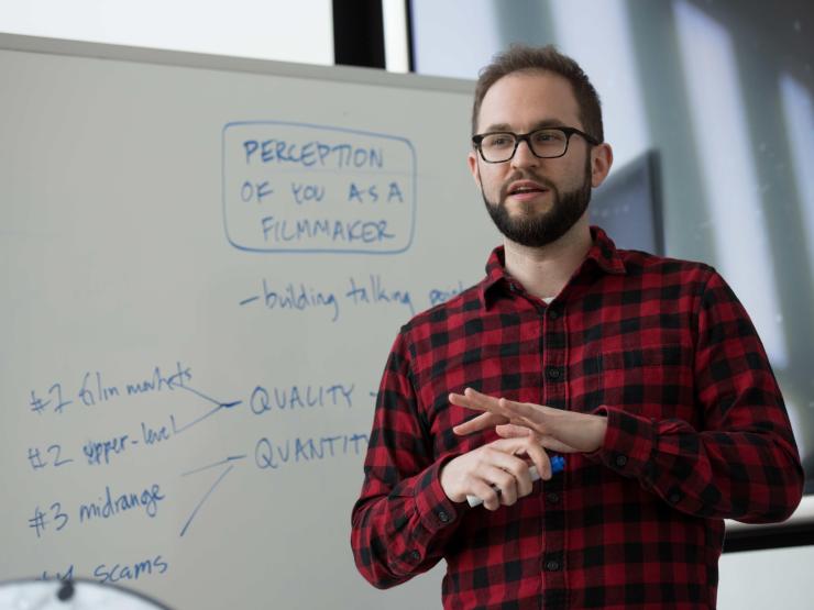 A man in a red shirt and glasses giving a speech in from of a whiteboard.