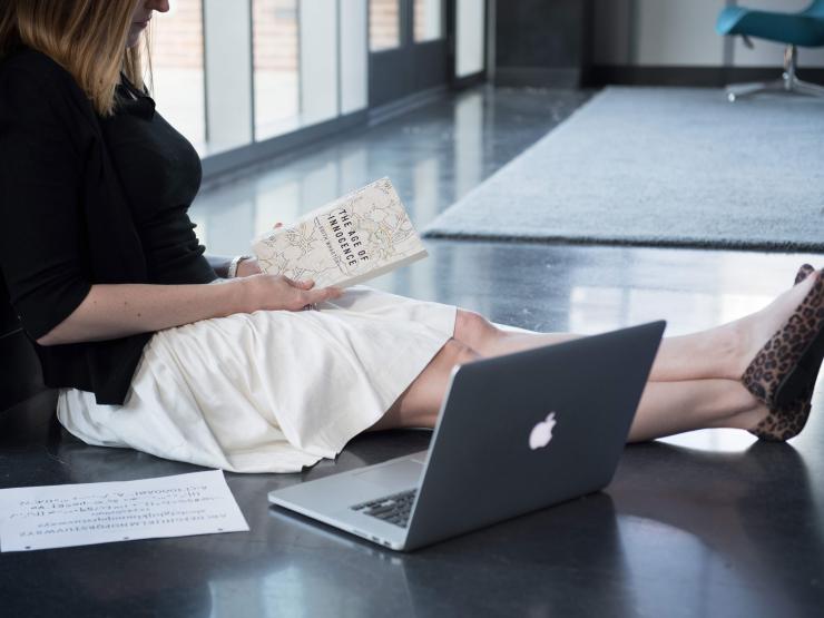 Student sitting on floor reading book next to laptop