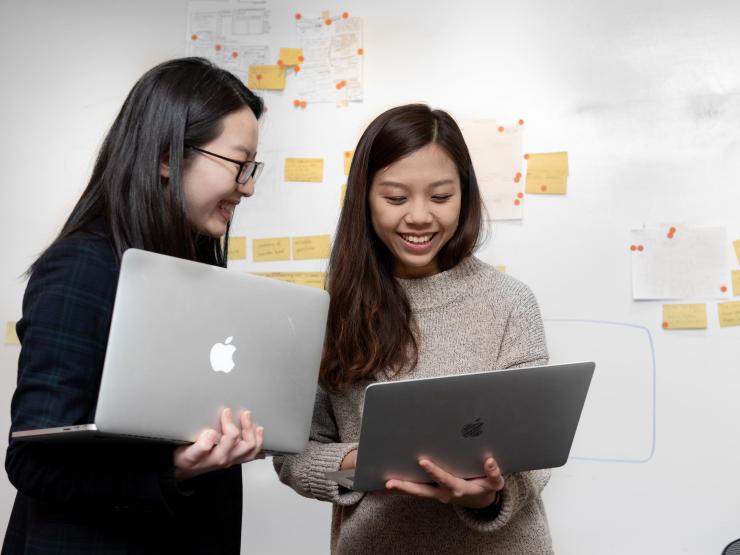 Two students looking at laptops with a whiteboard featuring post-it notes behind them