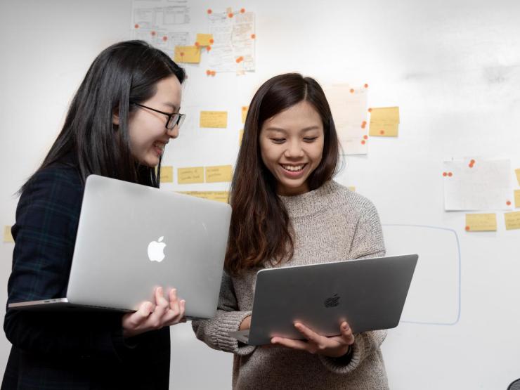 Two students smiling while using laptops in front of a dry erase board