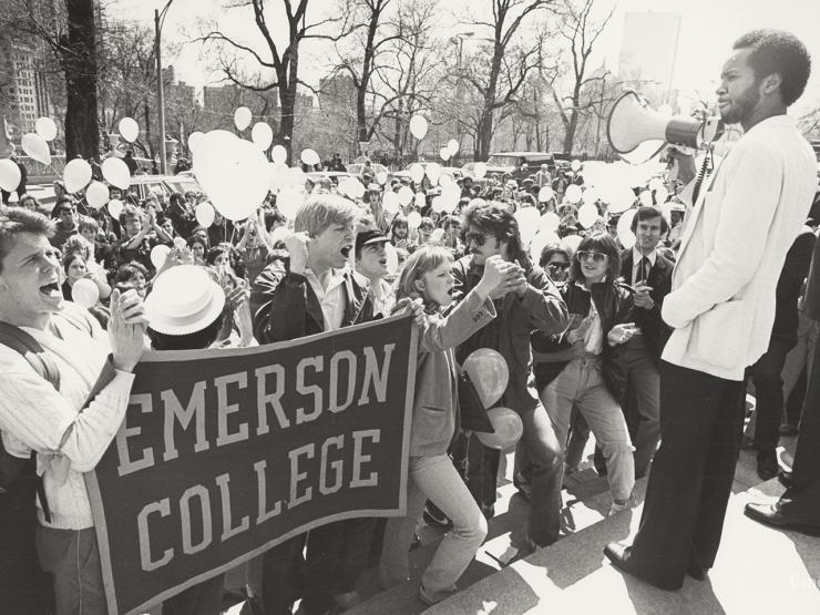 historical image of a crowd of students outside with balloons and a banner, in front of a stage