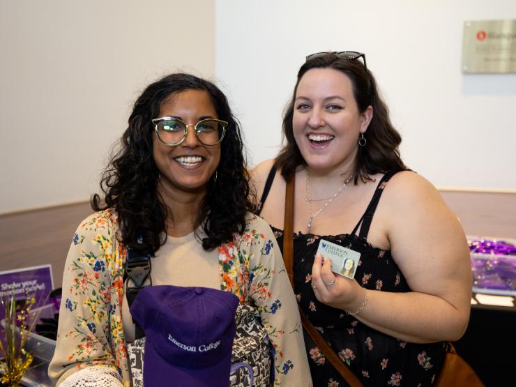 Two people standing next to each other and smiling at an Alumni event. They are standing near a table of swag for attendees. One is carrying a purple baseball cap, branded for Emerson College alumni.