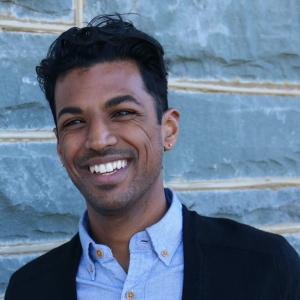 A man with brown skin, black hair, blue button up shirt, and black sweater smiling. 