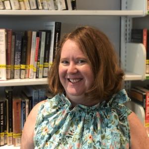 Cheryl McGrath, Executive Director Iwasaki Library and Learning, standing in front of a shelf of reserve books, wearing a top by Rachel Walters Collection. 