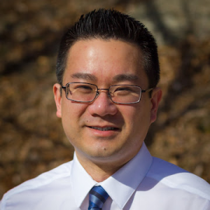 Chinese-American man with black hair and glasses wearing a white shirt and blue tie