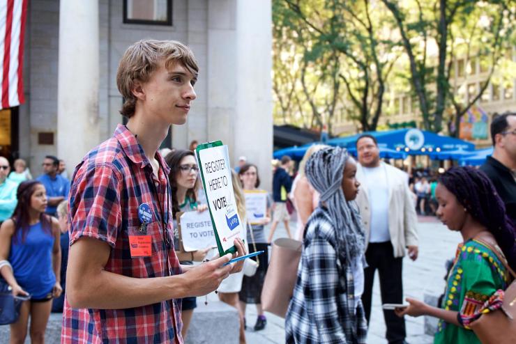 Young person at Faneuil Hall holding up a clipboard reading "Register Here to Vote"