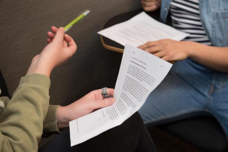 A pair of hands holding a piece of paper across from a student sitting at a desk. Both students are out of shot