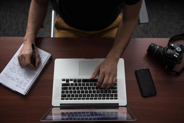 Overhead shot of a student typing on a MacBook with one hand and writing in a notebook with the other.